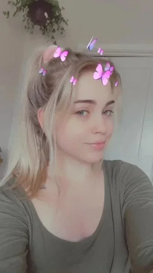 Woman with blonde hair in a ponytail, wearing a green top, taking a selfie indoors with butterfly
