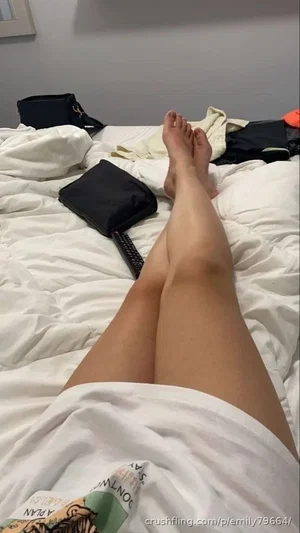 Person lying on bed, legs extended, wearing white shirt, bare legs visible, indoor setting.
