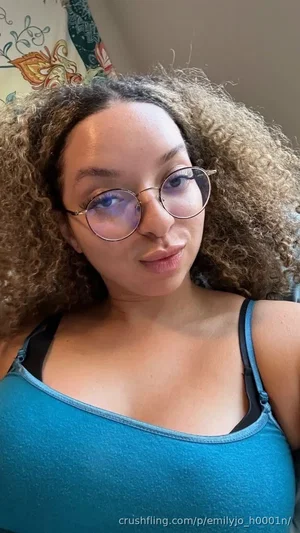 Woman with curly hair and glasses wearing a blue tank top indoors.