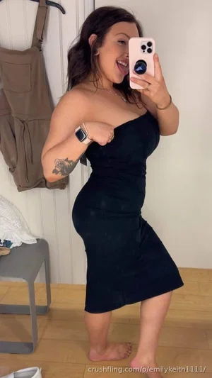 Woman in black one-shoulder dress posing for mirror selfie indoors, barefoot, tattoo on arm.