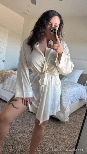 Woman in white robe taking mirror selfie in bedroom, tattoo on thigh.