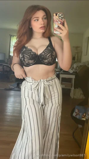 Woman with red hair in black lace bra and striped pants taking mirror selfie in bedroom.