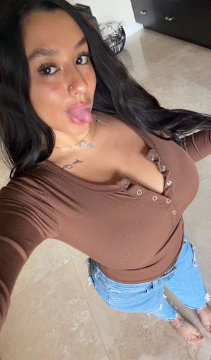 Curvy woman with long dark hair in brown top taking indoor selfie.