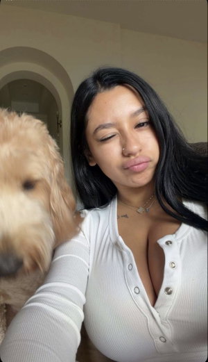 Woman with long black hair in white top taking selfie with dog indoors.