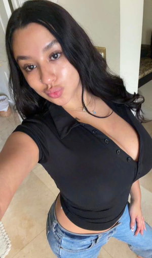 Woman with long dark hair in black top and jeans taking selfie indoors.