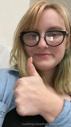 Blonde woman with glasses giving thumbs up, wearing denim shirt indoors.
