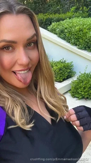 Woman with long blonde hair in black top sticking tongue out, outdoors with greenery.