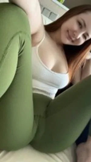 Woman with long brown hair in white tank top and green leggings posing indoors.