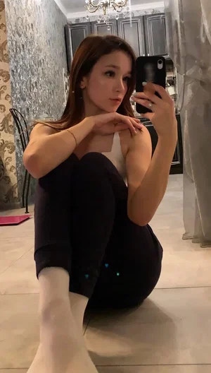 Woman in white top and black pants sits on floor, taking mirror selfie in decorated room.