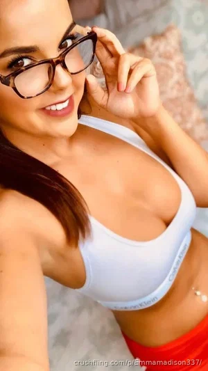 Woman in white bra and red bottoms, wearing glasses, smiling in a bedroom selfie.