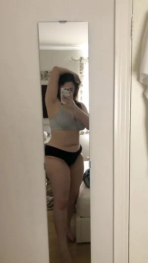 Curvy woman in gray bra, black panties, bedroom mirror selfie.