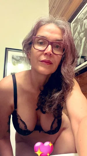 Woman with gray hair and glasses in black lace bra, indoors.