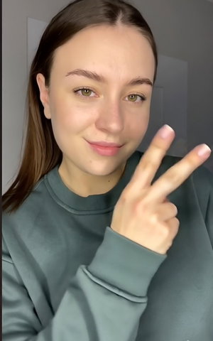 Slim woman with long brown hair in green hoodie making peace sign indoors.