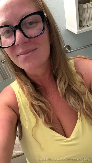 Woman with blonde hair and glasses in yellow top, taking selfie in bathroom.