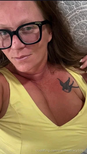 Woman with glasses and chest tattoo wearing yellow top, indoor selfie.