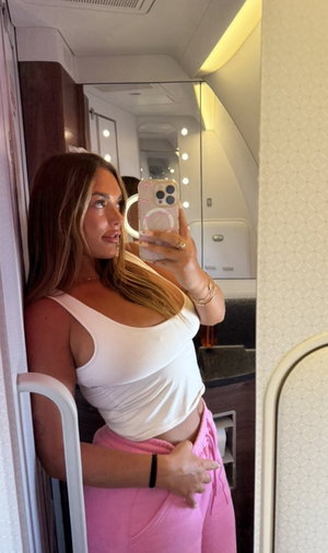 Woman taking mirror selfie in white top and pink pants on plane.