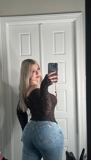 Woman with blonde hair in black lace top and jeans, taking mirror selfie indoors.