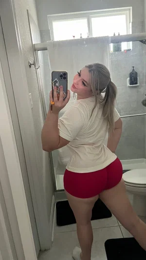 Woman with light hair in ponytail wearing white shirt and red shorts, taking mirror selfie in