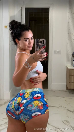Woman in white crop top and colorful shorts taking mirror selfie in indoor setting.