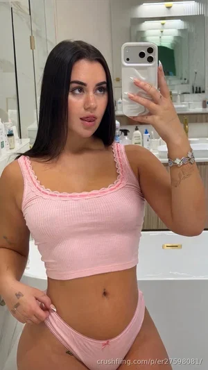 Woman in pink ribbed crop top and matching underwear, taking mirror selfie in bathroom.