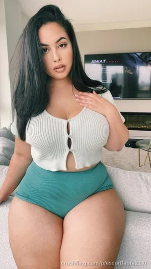 Woman with long dark hair in white ribbed top and teal shorts sitting on couch indoors.