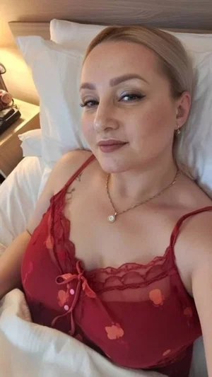 Woman with blonde hair in red floral lingerie lying in bed with white sheets.
