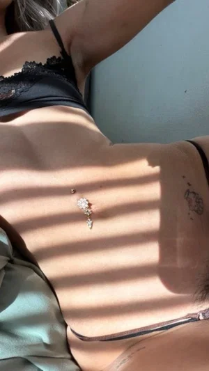 Woman in black lace lingerie lying on her stomach, back visible with navel piercing and tattoo.