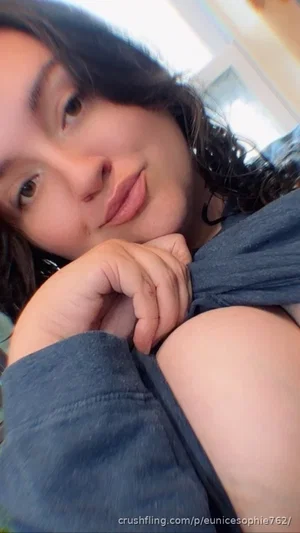 Woman with dark curly hair in a blue top, resting chin on hand, indoor setting.