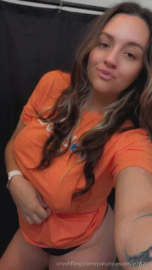 Woman with long wavy hair in orange t-shirt, posing in front of black backdrop.