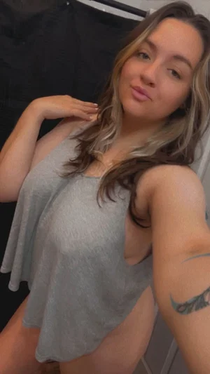 Woman with wavy hair in gray tank top, taking mirror selfie indoors.