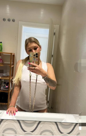 Blonde woman in white tank top taking mirror selfie in bathroom.