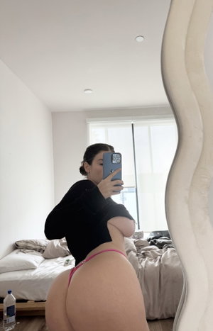 Woman in black top and pink underwear taking mirror selfie in bedroom.