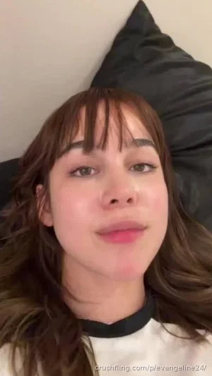 Woman with brown hair and bangs lying on bed, wearing white top with black collar.
