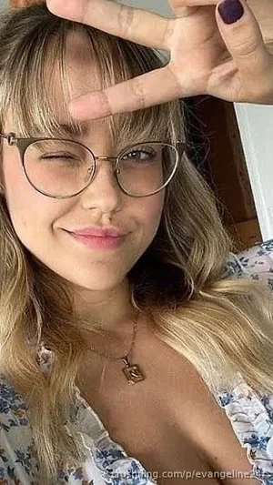 Woman with blonde hair and glasses, wearing a floral top, making a peace sign indoors.