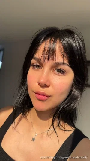 Woman with dark hair and bangs wearing black tank top, indoors, close-up selfie.