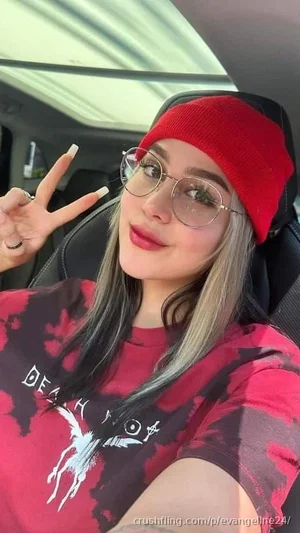 Woman with gray hair and red beanie in car, wearing red graphic tee, making peace sign.