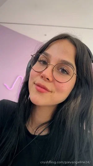 Woman with long dark hair and round glasses, wearing black top, indoors with pink wall and neon