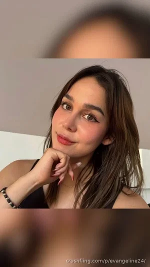 Woman with long brown hair in black top posing indoors, hand on chin.