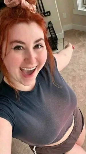 Woman with red hair in a bun, wearing a dark top and shorts, smiling in a home setting.