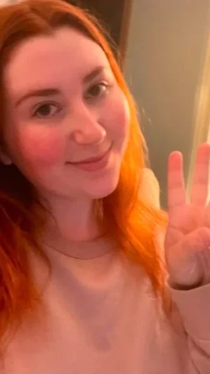 Woman with red hair in a light top waving at camera indoors.