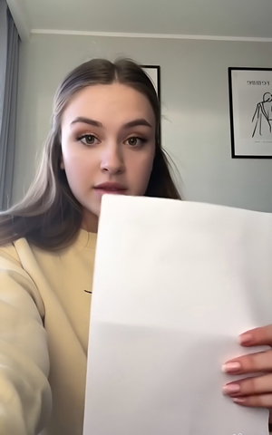 Petite woman with brown hair in beige sweatshirt holding paper indoors.