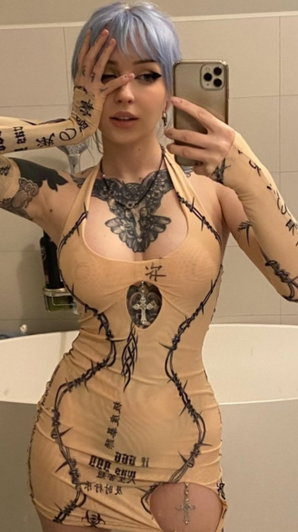 Woman with blue hair and tattoos wears a beige body suit with barbed wire print in a bathroom.