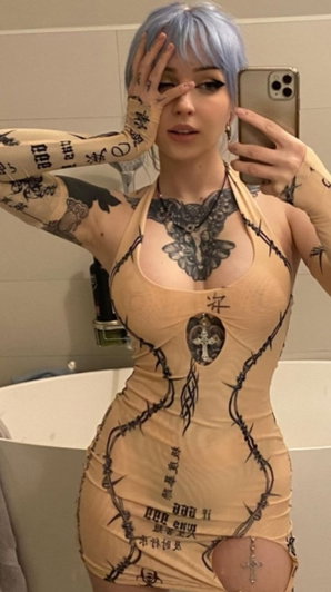 Woman with blue hair and tattoos wears a beige body suit with barbwire print, taking mirror selfie