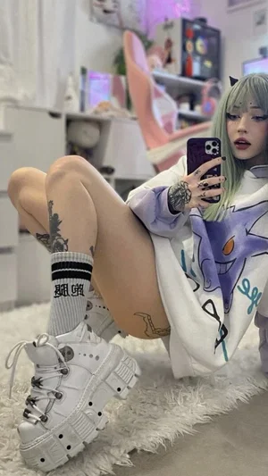 Woman with green hair and tattoos in oversized hoodie, sitting on fluffy rug, taking mirror selfie.