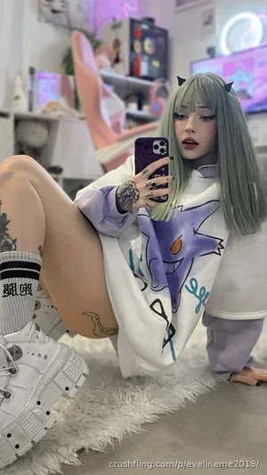 Woman with green hair and tattoos in oversized hoodie, taking mirror selfie in bedroom.