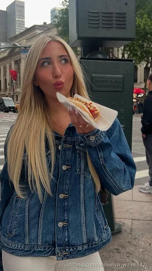 Blonde woman in denim jacket eating a hot dog on city street.