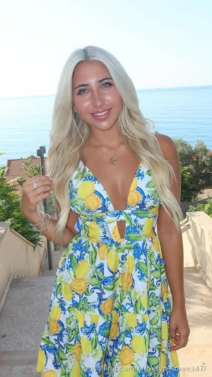 Woman with blonde hair in a lemon-print dress standing outdoors near the ocean.