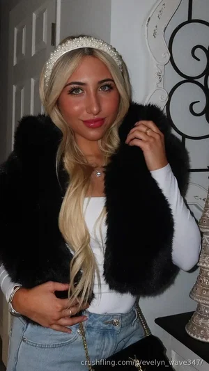 Blonde woman in white top, black fur vest, jeans, and pearl headband posing indoors.