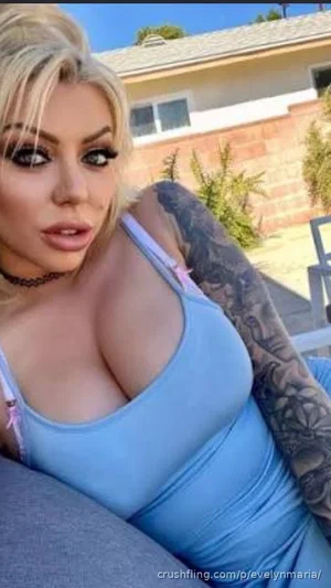 Woman with blonde hair and tattoos wears light blue tank top, sitting outdoors near a house.