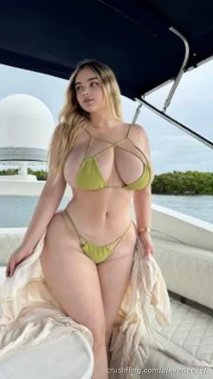Woman in green bikini posing on boat, blonde hair, curvy figure, outdoor setting.